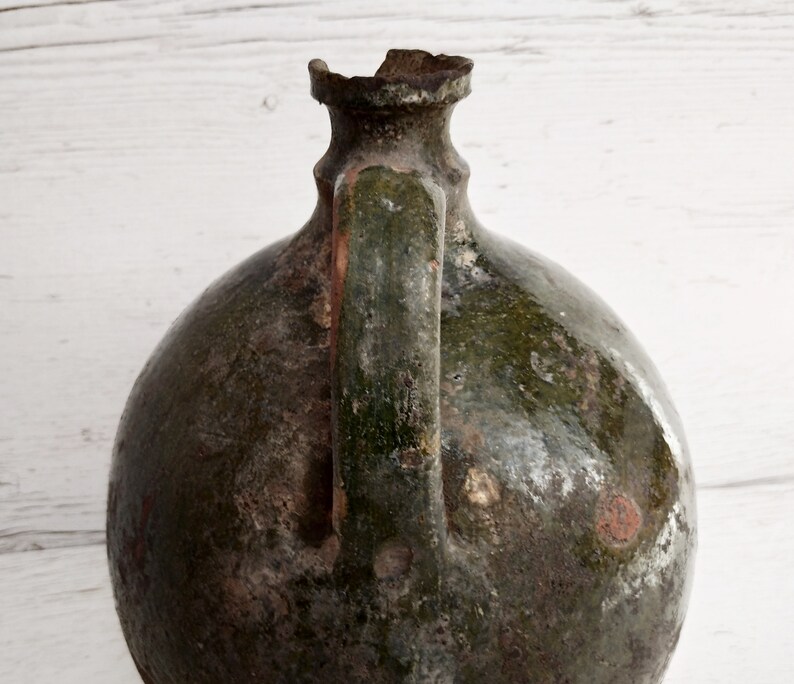 May include: A large, green, ceramic jug with a chipped rim and a handle. The jug has a rustic, aged appearance with a mottled green glaze and white spots.