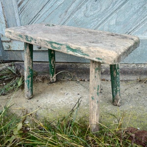 Puede incluir: Un taburete de madera r&uacute;stico con tres patas pintadas de verde. El taburete est&aacute; desgastado y muestra signos de antig&uuml;edad.