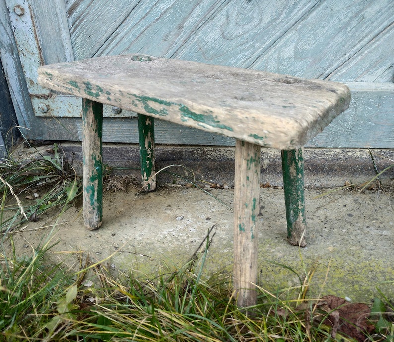 Puede incluir: Un taburete de madera r&uacute;stico con tres patas pintadas de verde. El taburete est&aacute; desgastado y muestra signos de antig&uuml;edad.