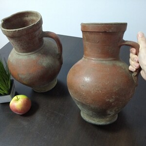 May include: Two brown terracotta pitchers with handles, one slightly tilted, on a dark wood surface with a small potted plant and a red apple.