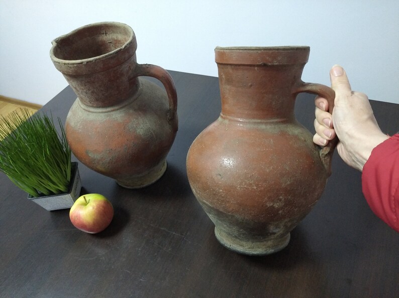 May include: Two brown terracotta pitchers with handles, one slightly tilted, on a dark wood surface with a small potted plant and a red apple.