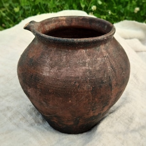May include: A brown clay pot with a wide mouth and a small spout. The pot has a rustic, aged appearance and is slightly chipped.