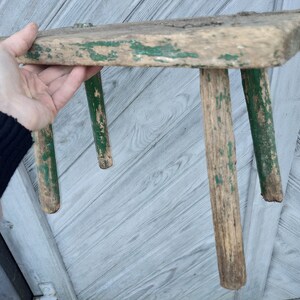 Puede incluir: Un taburete de madera r&uacute;stico con tres patas pintadas de verde. El taburete est&aacute; desgastado y muestra signos de antig&uuml;edad.