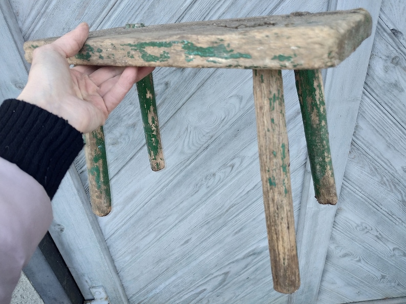 Puede incluir: Un taburete de madera r&uacute;stico con tres patas pintadas de verde. El taburete est&aacute; desgastado y muestra signos de antig&uuml;edad.