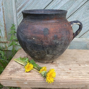 May include: A dark brown clay pot with a wide mouth and a single handle. The pot is sitting on a wooden surface with yellow dandelions.