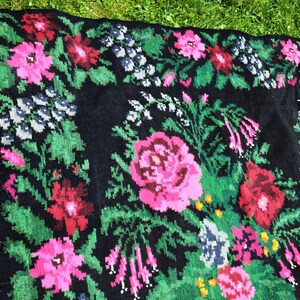 May include: A black wool rug with a floral pattern featuring pink, red, and green flowers. The rug has a fringe border.