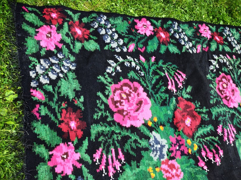 May include: A black wool rug with a floral pattern featuring pink, red, and green flowers. The rug has a fringe border.