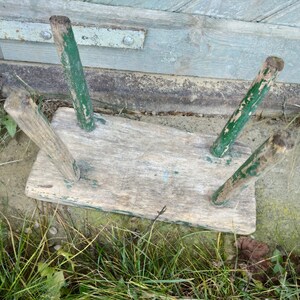 Puede incluir: Un taburete de madera r&uacute;stico con tres patas pintadas de verde. El taburete est&aacute; desgastado y tiene un acabado de madera natural.