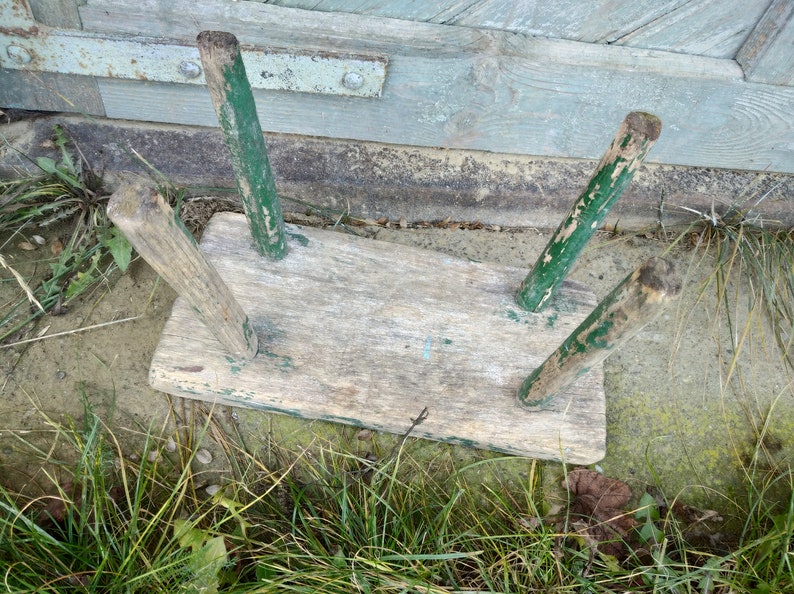 Puede incluir: Un taburete de madera r&uacute;stico con tres patas pintadas de verde. El taburete est&aacute; desgastado y tiene un acabado de madera natural.