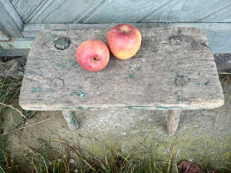 Puede incluir: Dos manzanas rojas sobre un taburete de madera r&uacute;stico con pintura verde. El taburete tiene dos patas y est&aacute; sentado sobre una superficie de tierra y hierba.