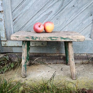 Puede incluir: Un taburete de madera r&uacute;stico con pintura verde, desgastado y descolorido, se encuentra sobre una superficie de hormig&oacute;n con hierba verde. Dos manzanas, una roja y una amarilla, descansan sobre la parte superior del taburete.