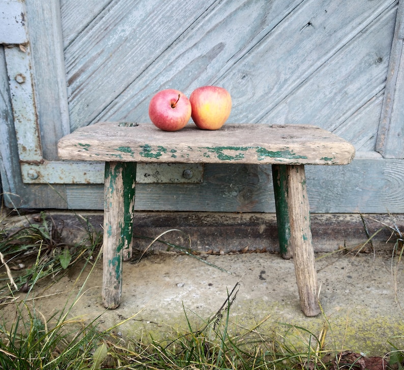 Puede incluir: Un taburete de madera r&uacute;stico con pintura verde, desgastado y descolorido, se encuentra sobre una superficie de hormig&oacute;n con hierba verde. Dos manzanas, una roja y una amarilla, descansan sobre la parte superior del taburete.