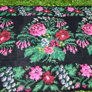May include: A black wool rug with a floral pattern. The rug features pink, red, green, and white flowers. The flowers are arranged in a symmetrical pattern.