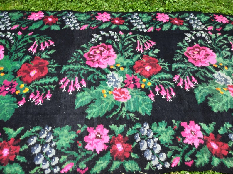 May include: A black wool rug with a floral pattern. The rug features pink, red, green, and white flowers. The flowers are arranged in a symmetrical pattern.