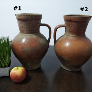 May include: Two brown ceramic jugs with handles, labeled #1 and #2. The jugs are on a dark brown wooden surface with a small potted plant and a red apple.