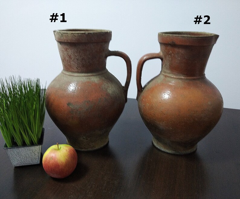 May include: Two brown ceramic jugs with handles, labeled #1 and #2. The jugs are on a dark brown wooden surface with a small potted plant and a red apple.