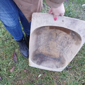May include: A large, wooden, oval-shaped bowl with a worn, rustic finish. The bowl is held by a person's hand, and the bowl is resting on a bed of green grass.