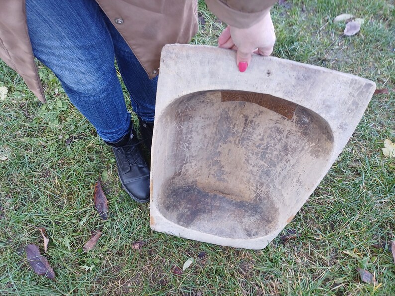 May include: A large, wooden, oval-shaped bowl with a worn, rustic finish. The bowl is held by a person's hand, and the bowl is resting on a bed of green grass.