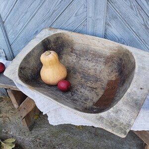 May include: A large, wooden, oval-shaped bowl with a rustic, weathered finish. The bowl is filled with a small, orange squash and three red apples.