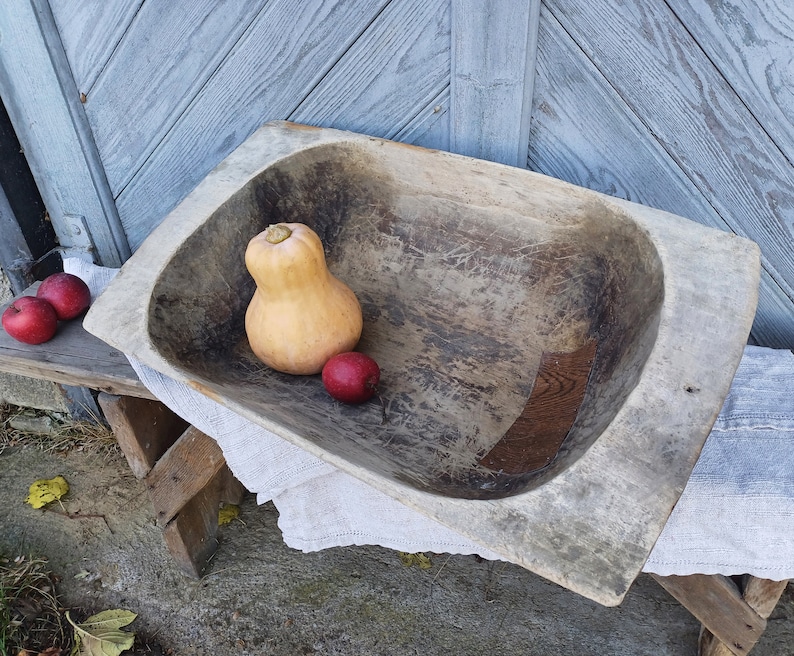 May include: A large, wooden, oval-shaped bowl with a rustic, weathered finish. The bowl is filled with a small, orange squash and three red apples.