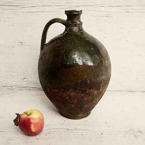 May include: A large, green and brown ceramic jug with a handle. The jug has a rustic, aged appearance. A red apple sits in front of the jug.