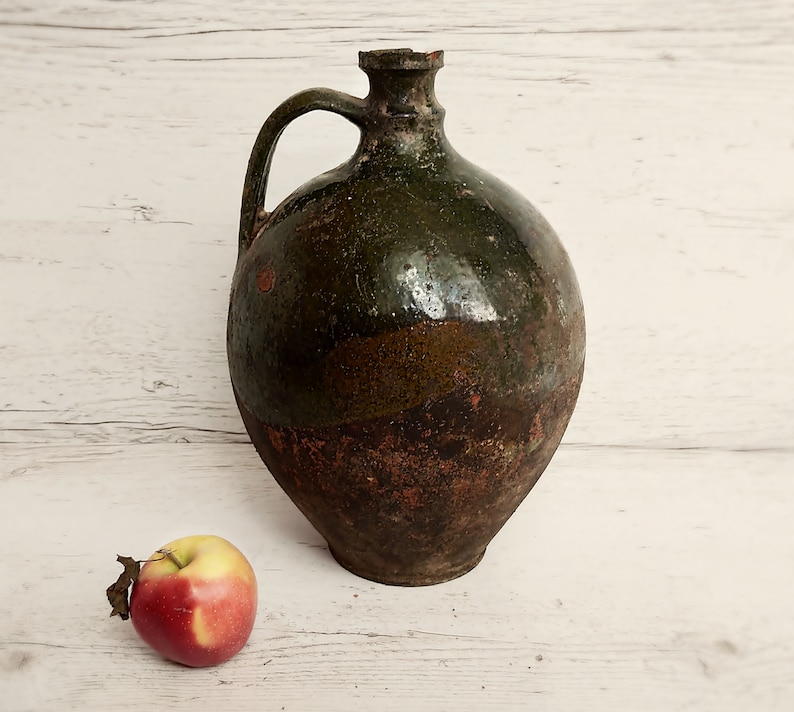 May include: A large, green and brown ceramic jug with a handle. The jug has a rustic, aged appearance. A red apple sits in front of the jug.