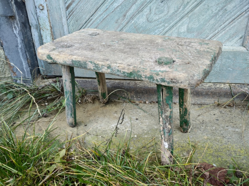 Puede incluir: Un taburete de madera r&uacute;stico con un acabado de pintura verde desgastada. El taburete tiene cuatro patas y una parte superior rectangular.