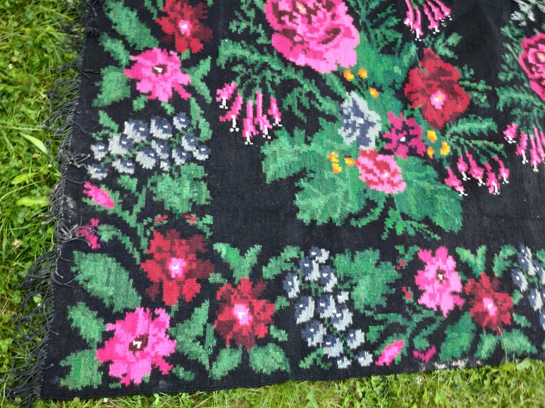 May include: A black rug with a floral pattern. The rug features pink, red, and green flowers on a black background. The rug has a fringe border.
