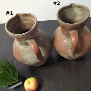 May include: Two terracotta pitchers with handles, labeled #1 and #2, on a wooden surface. The pitchers are dusty and have a rustic appearance.