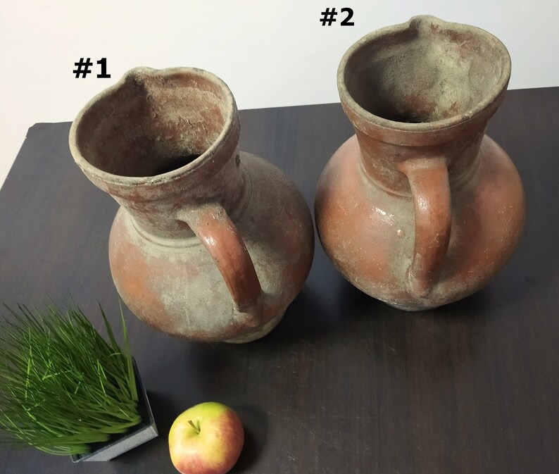 May include: Two terracotta pitchers with handles, labeled #1 and #2, on a wooden surface. The pitchers are dusty and have a rustic appearance.