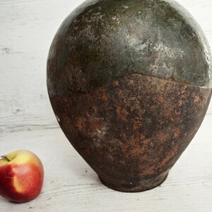 May include: A large, round, ceramic jar with a green glaze on the top half and a brown glaze on the bottom half. The jar is sitting on a white wooden surface. A red apple is in the foreground.