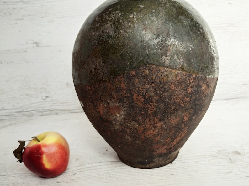 May include: A large, round, ceramic jar with a green glaze on the top half and a brown glaze on the bottom half. The jar is sitting on a white wooden surface. A red apple is in the foreground.