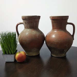 May include: Two terracotta pitchers with a rustic, aged look. The pitchers have a wide base and a narrow neck, with a handle on one side. They are sitting on a dark wooden surface.