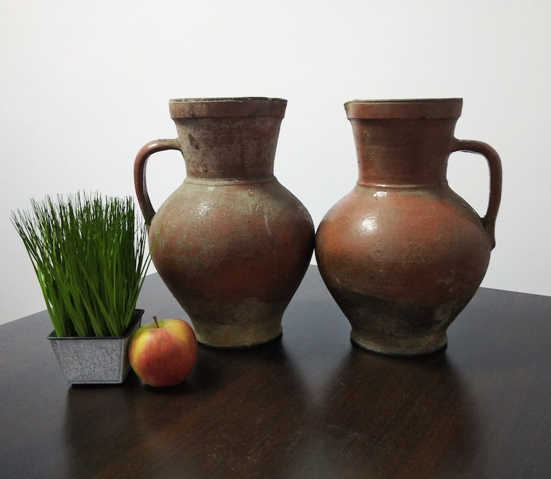 May include: Two terracotta pitchers with a rustic, aged look. The pitchers have a wide base and a narrow neck, with a handle on one side. They are sitting on a dark wooden surface.