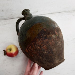 May include: A large, round, green and brown ceramic jug with a handle. The jug has a rustic, aged appearance.