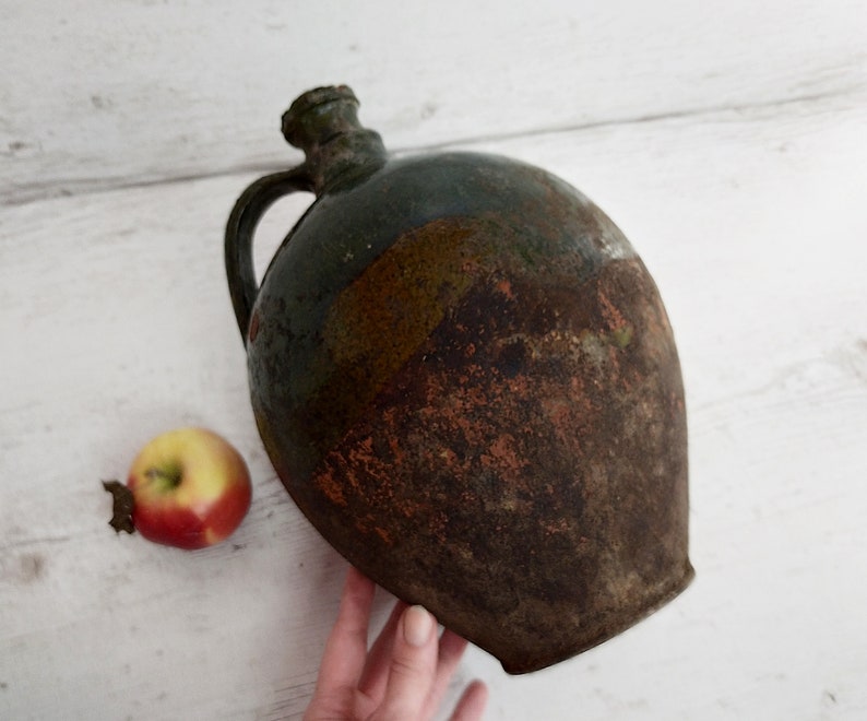 May include: A large, round, green and brown ceramic jug with a handle. The jug has a rustic, aged appearance.