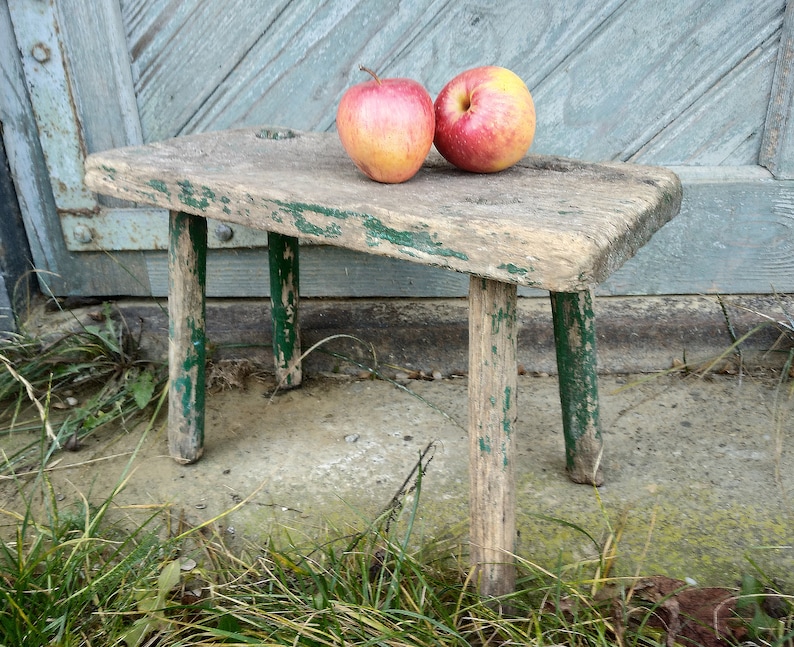 Puede incluir: Un taburete de madera r&uacute;stico con pintura verde, desgastado y descascarado, se encuentra sobre una superficie de hormig&oacute;n con hierba verde. Dos manzanas rojas est&aacute;n encima del taburete.