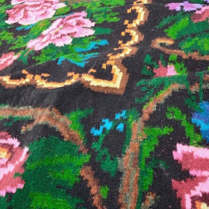 May include: Close-up of a colourful woven rug with a floral pattern. The rug is made of wool and has a black background with pink, green, blue, and orange flowers.