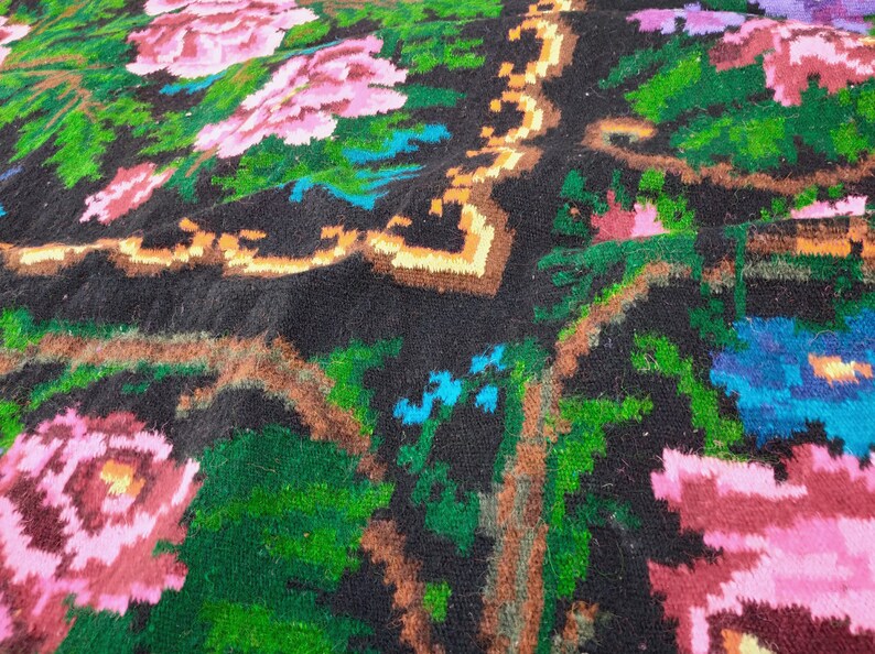 May include: Close-up of a colourful woven rug with a floral pattern. The rug is made of wool and has a black background with pink, green, blue, and orange flowers.