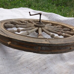 May include: A large wooden spinning wheel with a metal crank. The wheel is made of light brown wood and has a dark brown rim. The spokes are made of a lighter wood and have a rounded knob at the end. The wheel is in a worn condition, with some of the wood showing signs of age and wear.