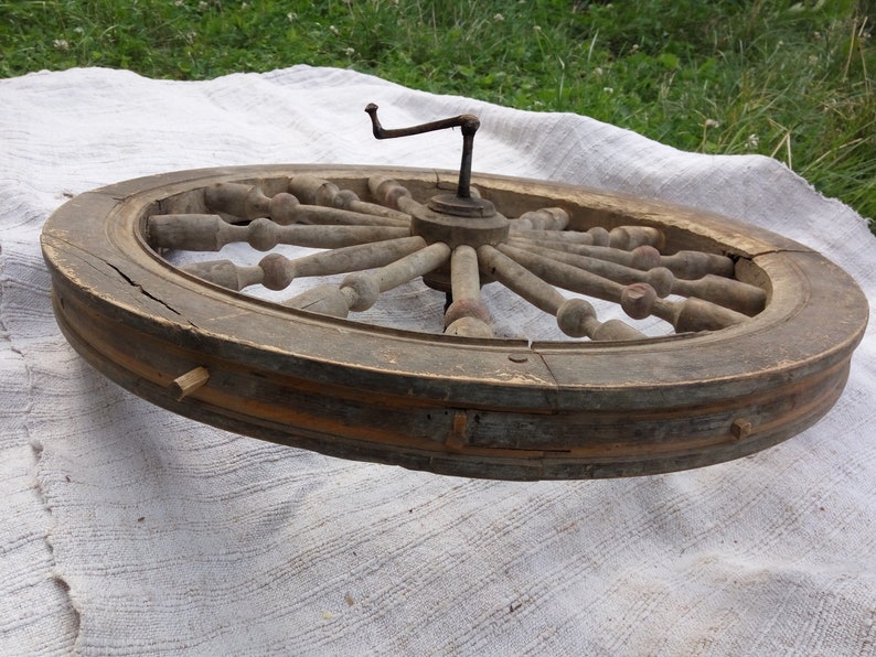 May include: A large wooden spinning wheel with a metal crank. The wheel is made of light brown wood and has a dark brown rim. The spokes are made of a lighter wood and have a rounded knob at the end. The wheel is in a worn condition, with some of the wood showing signs of age and wear.