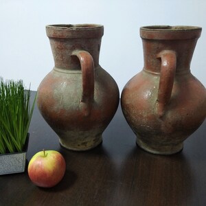 May include: Two terracotta pottery jugs with handles, one on the left and one on the right, on a dark wood surface. A small potted plant and a red apple are in the foreground.