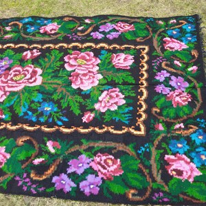 May include: A black wool rug with a floral pattern. The rug features pink, purple, blue, and green flowers on a black background. The rug has a fringed border.