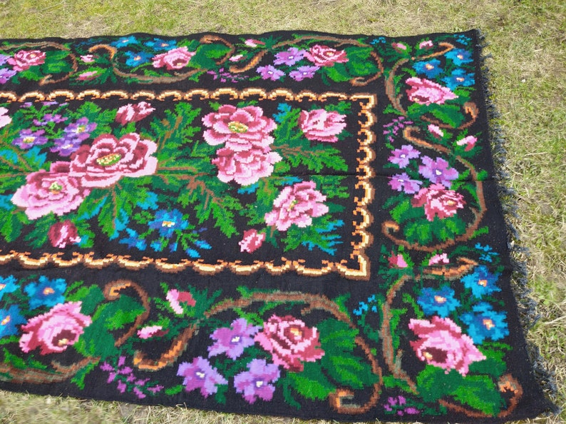 May include: A black wool rug with a floral pattern. The rug features pink, purple, blue, and green flowers on a black background. The rug has a fringed border.