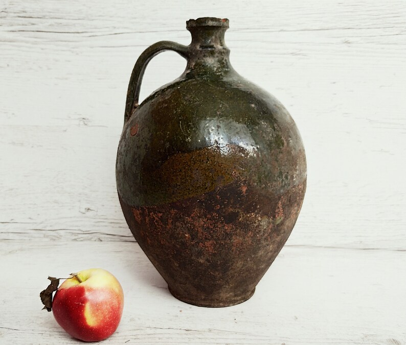 May include: A large, green and brown ceramic jug with a handle. The jug has a rustic, aged appearance.