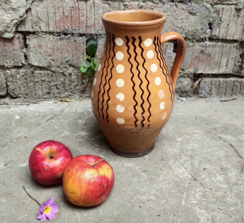 May include: A brown ceramic pitcher with a handle, decorated with white dots and black wavy lines. Two red apples with yellow accents and a small purple flower rest on a concrete surface near the pitcher.