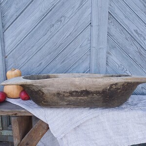 May include: A large, rustic wooden bowl with a dark brown finish. The bowl is sitting on a white linen cloth and a wooden table. There are several small red apples and a large orange pumpkin in the background.