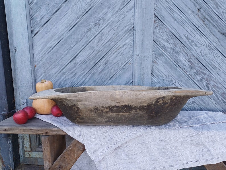 May include: A large, rustic wooden bowl with a dark brown finish. The bowl is sitting on a white linen cloth and a wooden table. There are several small red apples and a large orange pumpkin in the background.