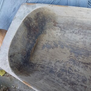 May include: A large, wooden dough bowl with a rectangular shape and a rounded bottom. The bowl is made of light-colored wood and has a worn, rustic appearance. The surface of the bowl is covered in scratches and grooves, indicating that it has been used extensively.
