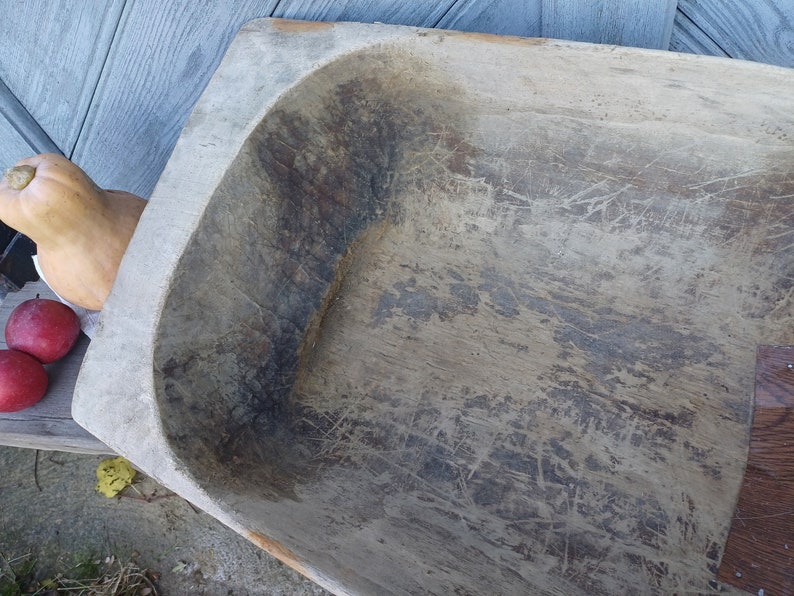 May include: A large, wooden dough bowl with a rectangular shape and a rounded bottom. The bowl is made of light-colored wood and has a worn, rustic appearance. The surface of the bowl is covered in scratches and grooves, indicating that it has been used extensively.